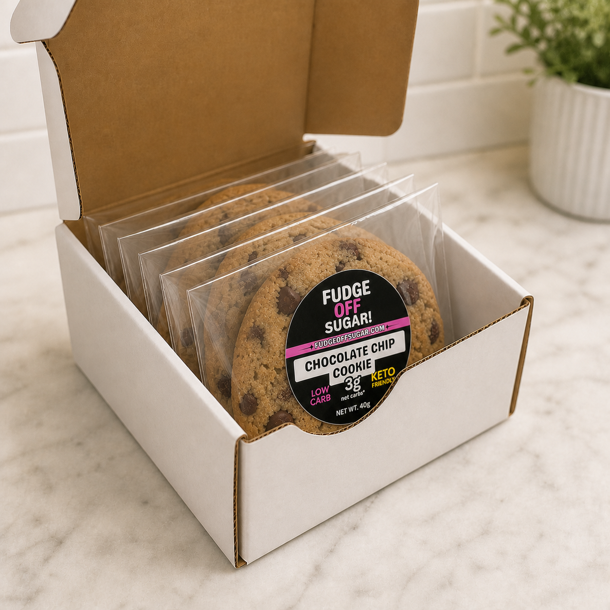 


A white cardboard box with the lid open, holding several individually wrapped chocolate chip cookies in clear plastic bags. The front cookie has a round black label reading 'FUDGE OFF SUGAR! KETO CHOCOLATE CHIP COOKIE'.